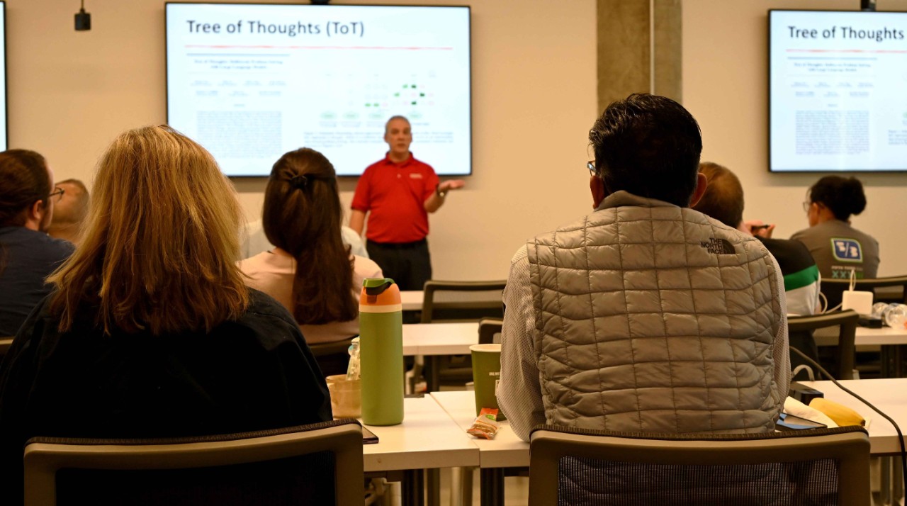 A man in a red shirt and and black pants instructs a class in front of a screen.