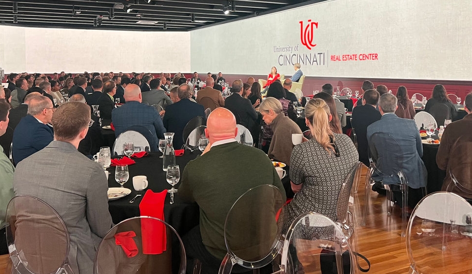 Attendees at a Real Estate Center Roundtable event sit at tables listening to a speaker standing on a stage at a podium with two screens to either side of them