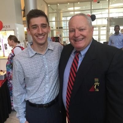 A man in a blue shirt and a man in a blue shirt and black jacket smile.