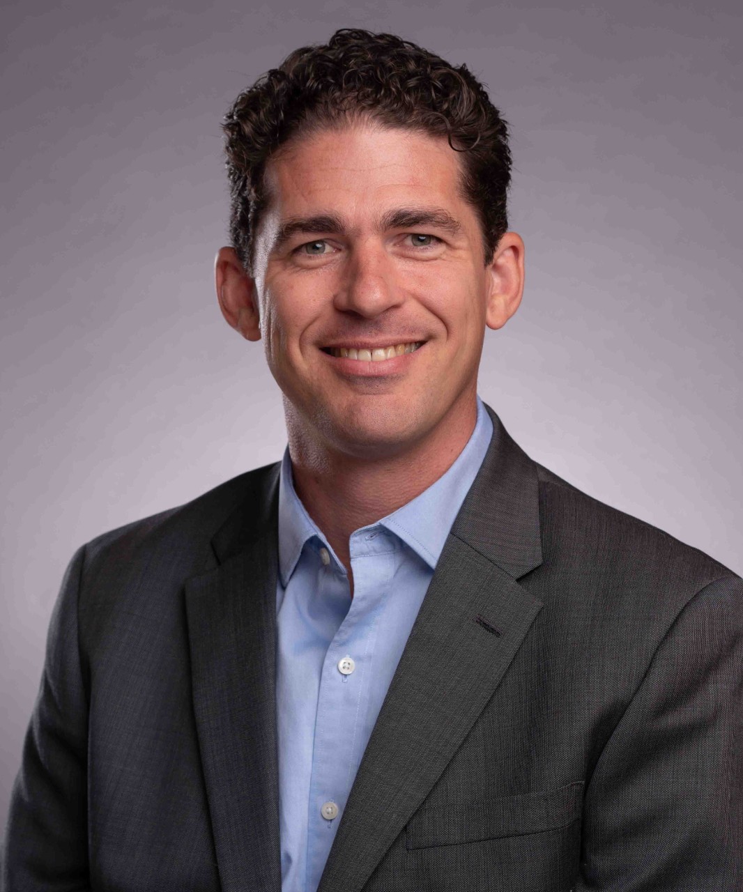 A man in a blue shirt and a dark blue jacket smiles.