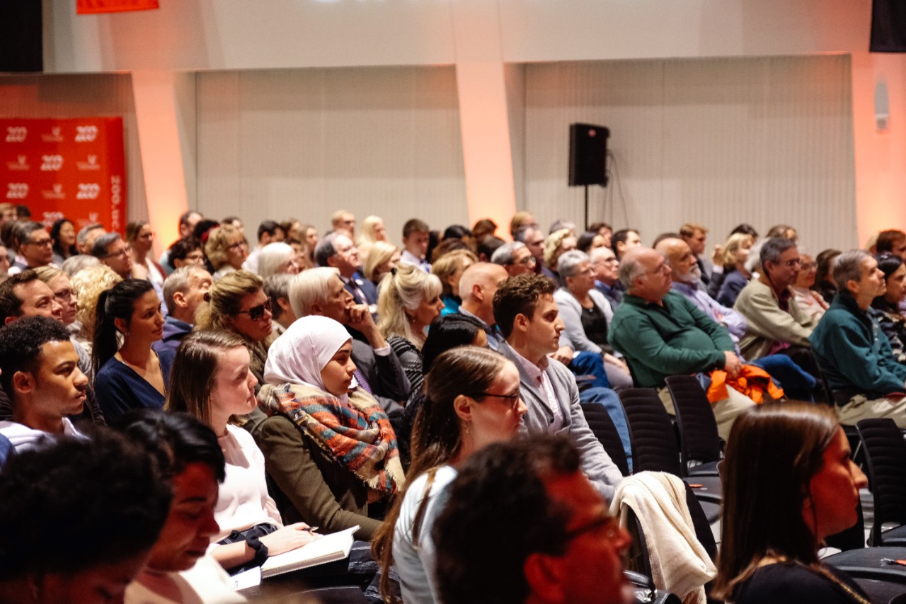 A crowd watches a person speak at a conference.