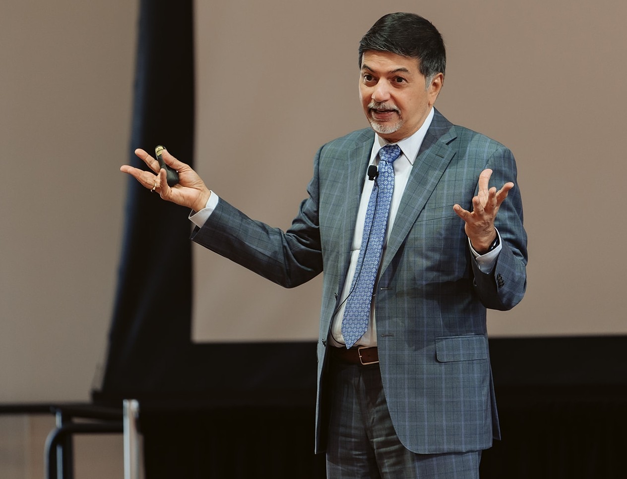 A man in a gray suit stands on a stage and speaks to a crowd.