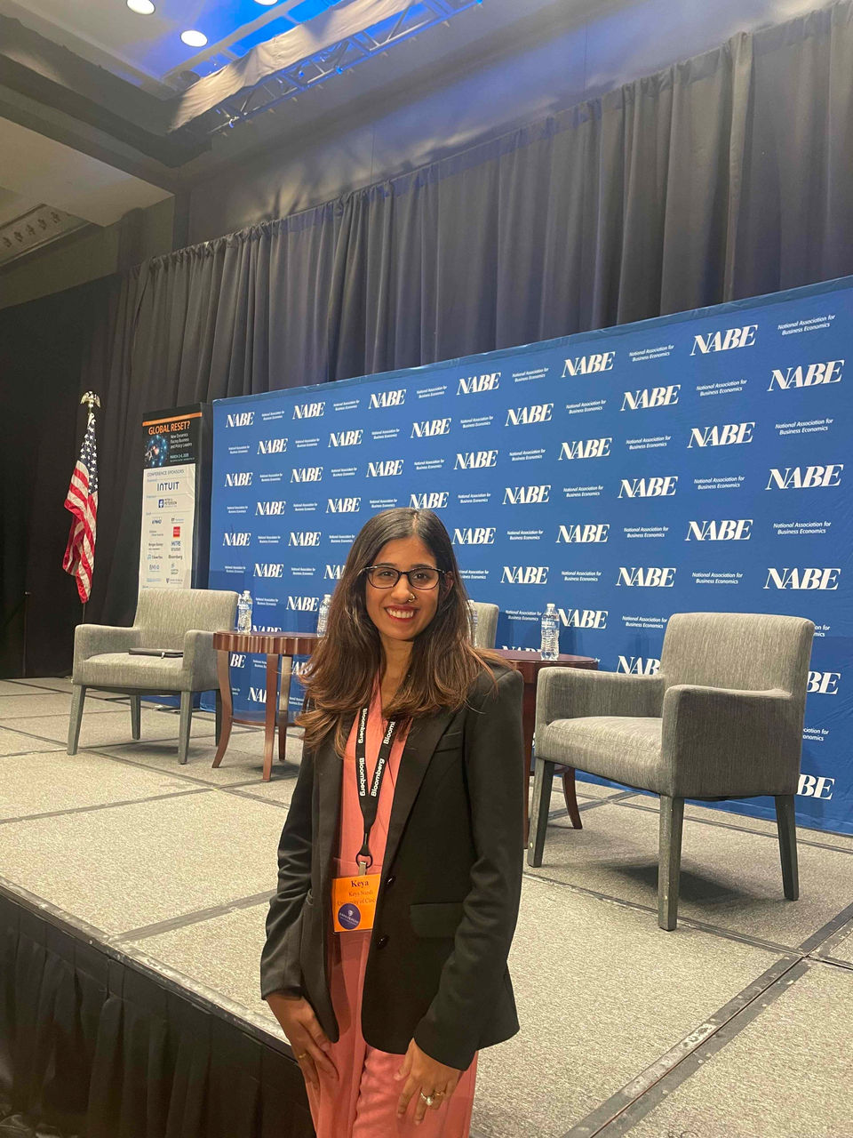 A female students in an orange dress and black jacket stands in front of a podium.