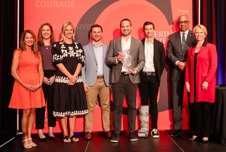 Men and women stand in front of a red banner while holding an award.