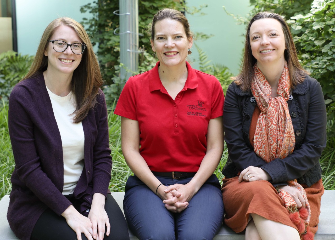 Three women in professional dress sit on a bench and smile.