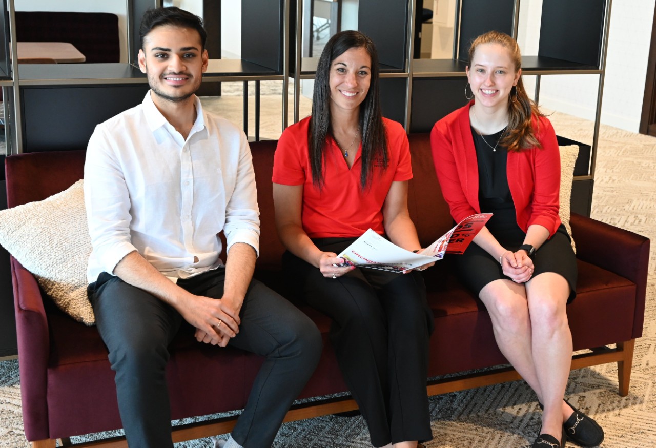 One man and two women in various colors of professional dress, seated with a woman holding a magazine.