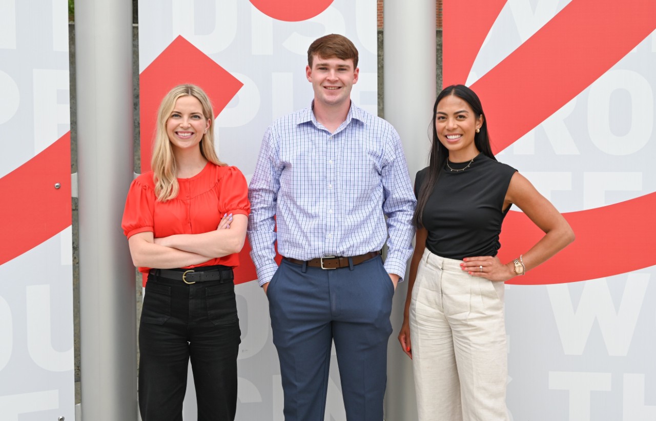Two women and one man in various professional dress stand outside in front of a sign.