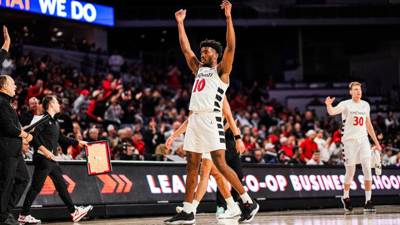 A basketball player in a white jersey walks off the court with a scoreboard and crowd in the background.