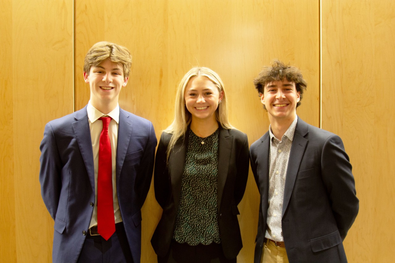 Three Lindner students in blazers pose in front of a wooden background