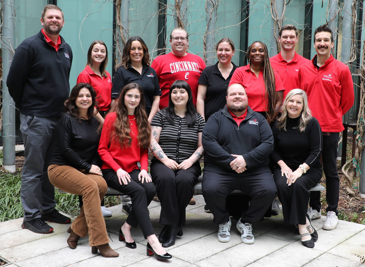 A group of men and women stand and sit in a courtyard, wearing red, black and white shirts and professional dress.