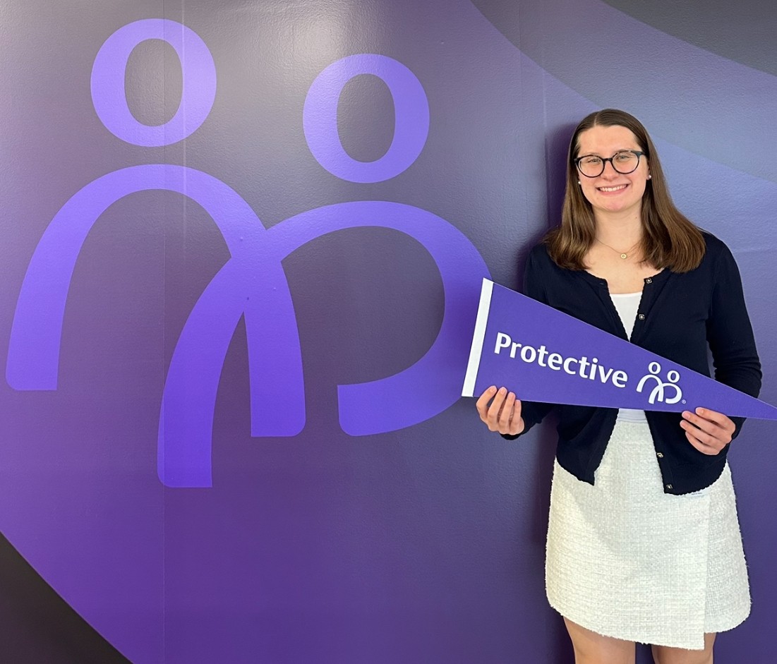 A student on co-op points hold a purple pennant and stands in front of a purple wall.