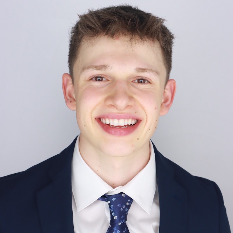 Headshot of Alex Davey in a white button-up shirt and navy jacket in front of grey lit background