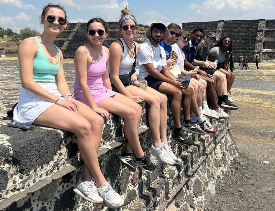A group of Lindner Business Honors students sit on a wall with a historic building in the background.