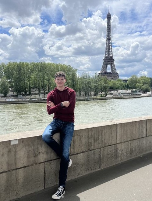 UC student posing with arms and legs crossed in front of a river with the Eiffel Tower in the background.